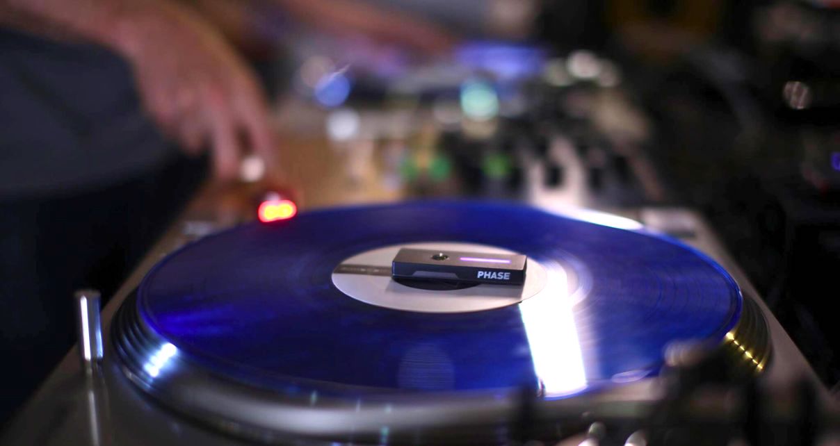 A DJ mixes on two turntables with blue records. There is a tiny Phase device on top of the vinyl in the foreground. 