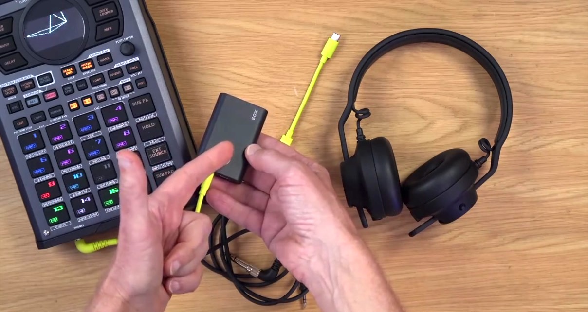 A Roland sampler and AIAIAI TMA-2 DJ headphones lay flat on a wooden desk. A person holds and points to a tiny box in their hands. 
