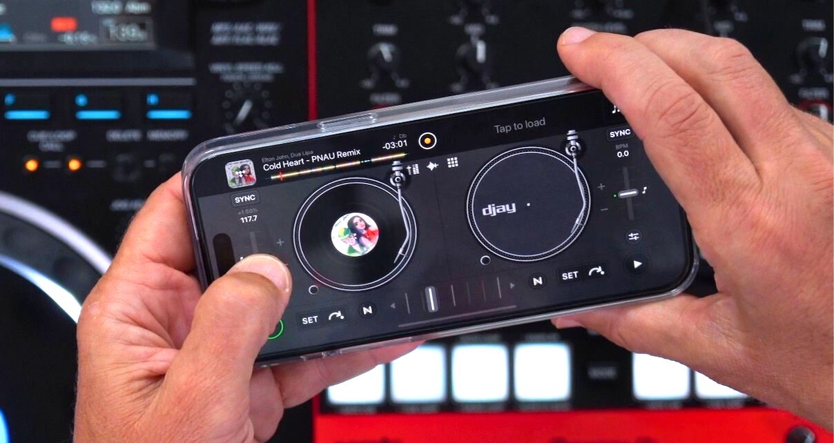 Top-down view of a mobile phone turned sideways, being held with both hands over a DJ set-up. On the screen is Djay Pro software. 