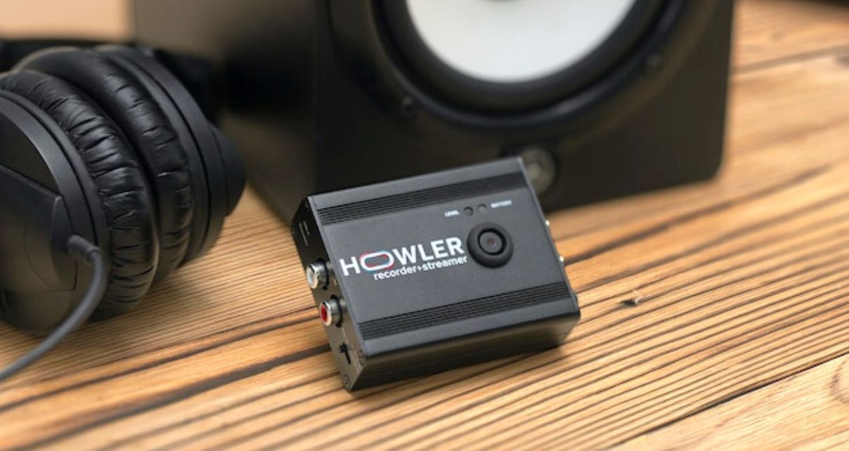 Small black Howler audio interface sat on a wooden desk in front of black headphones and a black and white monitor