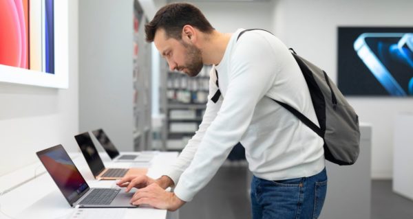A white man with short brown hair and a beard leans towards a laptop display in a store. He's wearing a long-sleeve white shirt, blue jeans, and a grey backpack. He seems to be considering a purchase as he types on a silver laptop. 
