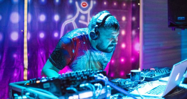 A white male DJ with dark hair and beard stands behind a booth. He's wearing headphones and bending over to squint at his laptop. In front of him are various pieces of DJ gear. Behind him is a colourful tapestry. 