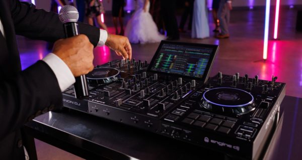 A DJ in a fancy suit stands behind a Prime 4+ controller. They're holding a microphone ready to talk while a wedding party dances in the background.