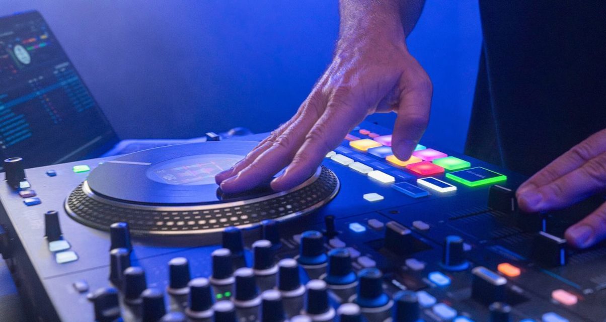 Angled view of a DJ touching the platters on a Rane Performer controller. In the background you can see a laptop displaying Serato DJ Pro software. The whole photo is lit in dark blue lighting.
