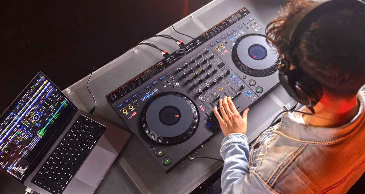 Birds eye view of a masculine-presenting brunette person standing behind a DJ booth, wearing black headphones and a denim jacket. They're looking down at an AlphaTheta DDJ-GRV6 controller and tweaking the volume levels. On the left is a silver MacBook Pro laptop displaying Rekordbox DJ software.