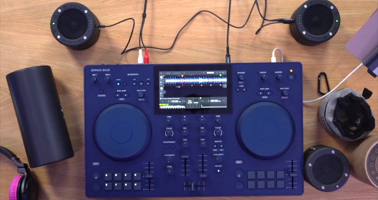 A top-down view of DJ gear set up on a wooden table. There are several black Minirig speakers and a subwoofer surrounding a deep blue AlphaTheta Omnis Duo standalone DJ system. 