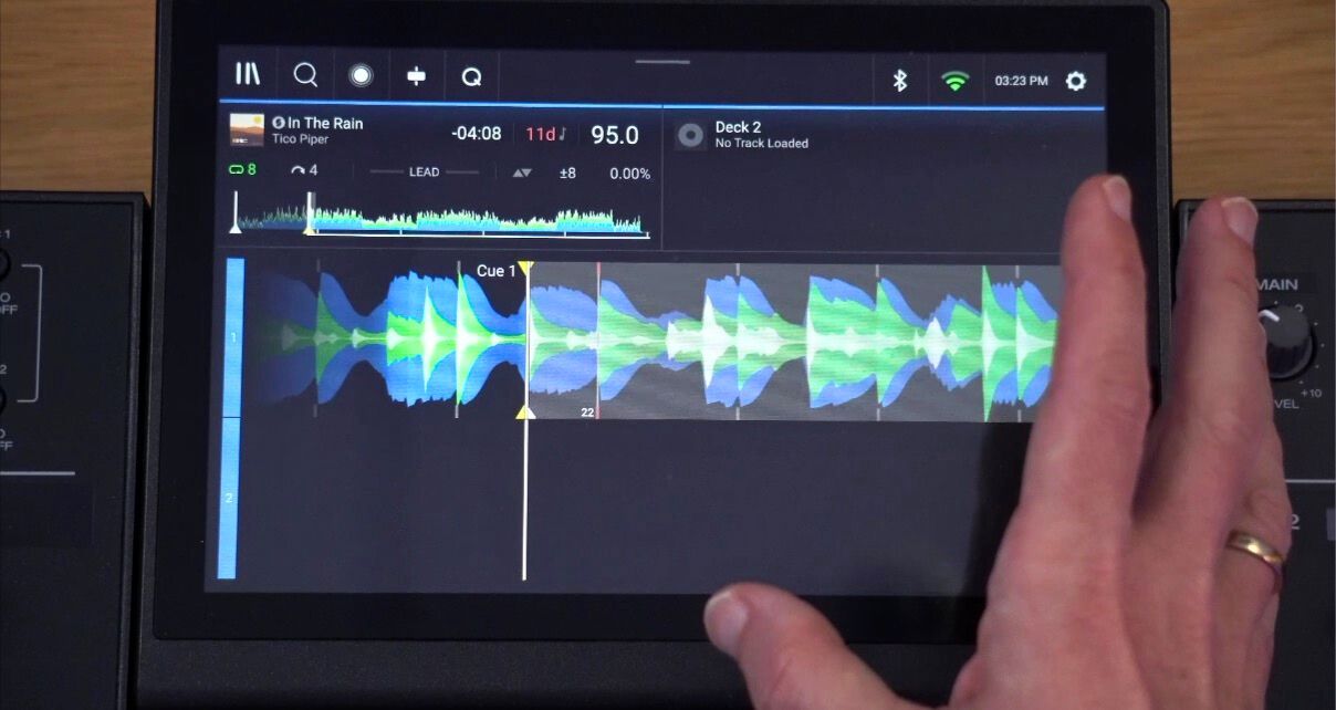Top-down view of the Denon DJ Prime 4+ screen laying on a wooden desk, with the hand of a white man gesturing towards the blue white and green track waveform