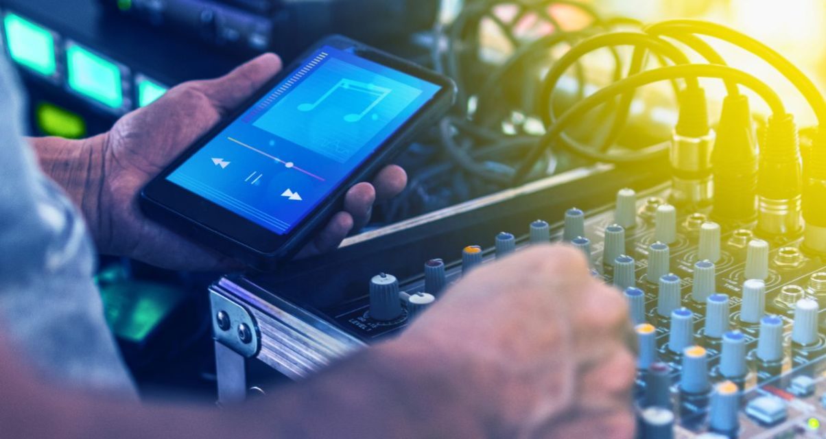 A white man holds a smart phone. The phone is in his left hand and displays a generic music app. With his right hand, he is tweaking various colourful knobs on a live sound mixer.