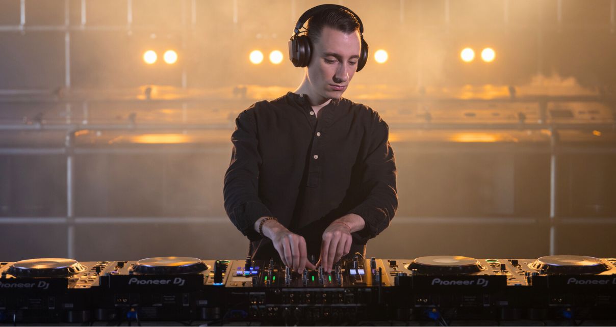 White male DJ with black headphones stands in a warmly lit room behind four CDJs and a mixer