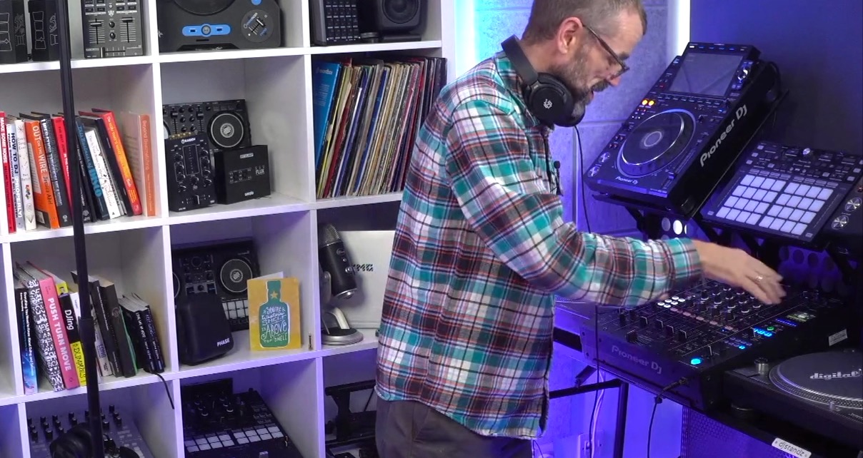 A white man with glasses, black headphones, and a flannel shirt stands in a DJ studio next to some club gear. 