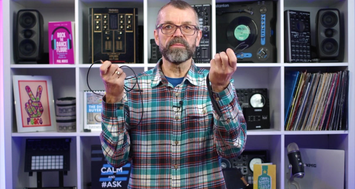 A white man with a beard, glasses, and a flannel shirt stands in front of white bookshelves holding two different types of cables, one in each hand. 