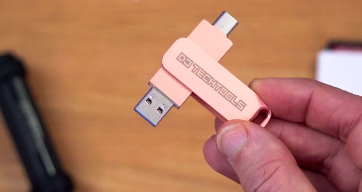 The hand of a white man holds a rose gold USB drive that has a DJ TechTools logo printed on it. In the background is a blurry wooden desk holding a black USB stick and a white SD card.