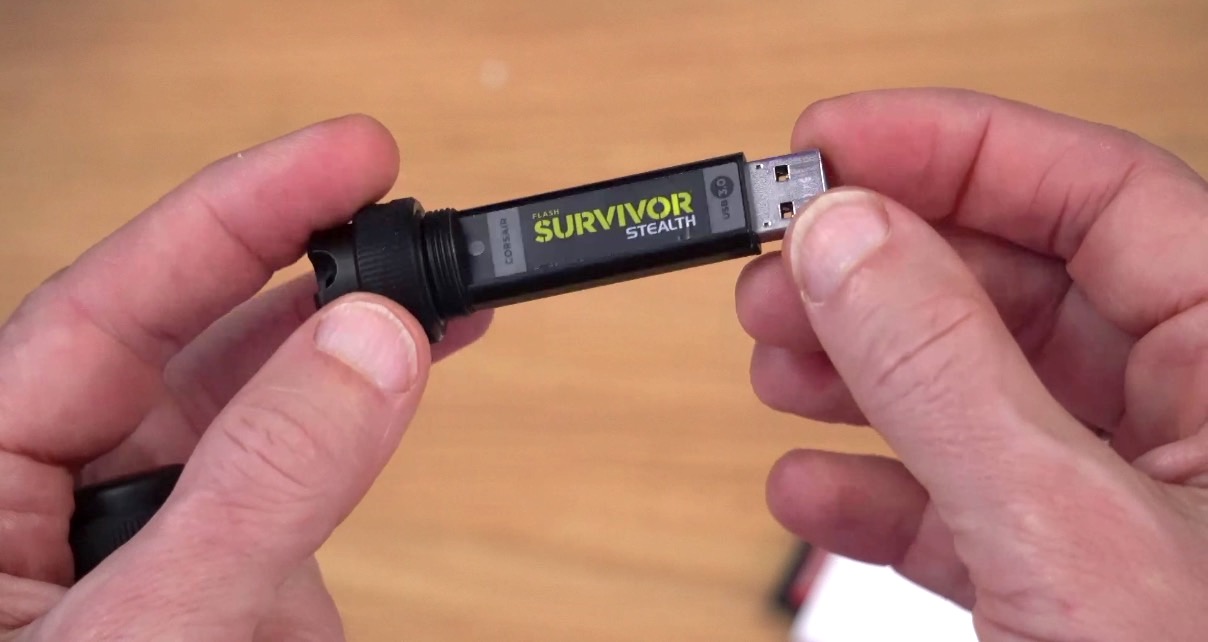 White person holding a black Corsair Survivor Stealth USB drive against the backdrop of a wooden desk.