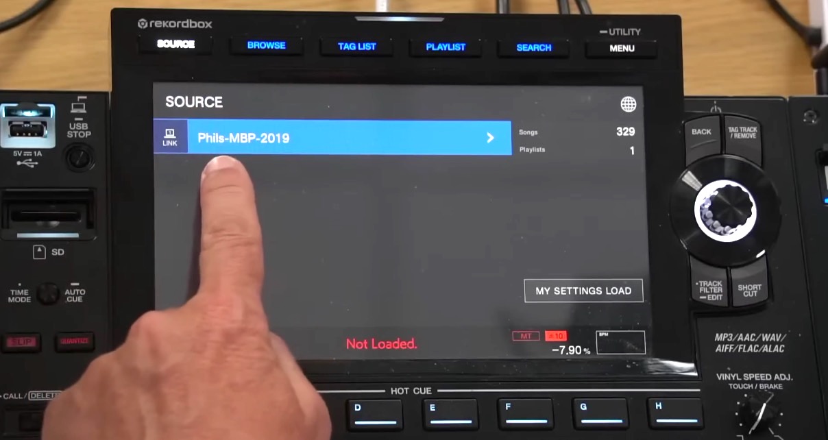 A white person points with their left hand at a standalone DJ gear screen that says the word "Source" at the top. Underneath that it says Phils-MBP-2019.