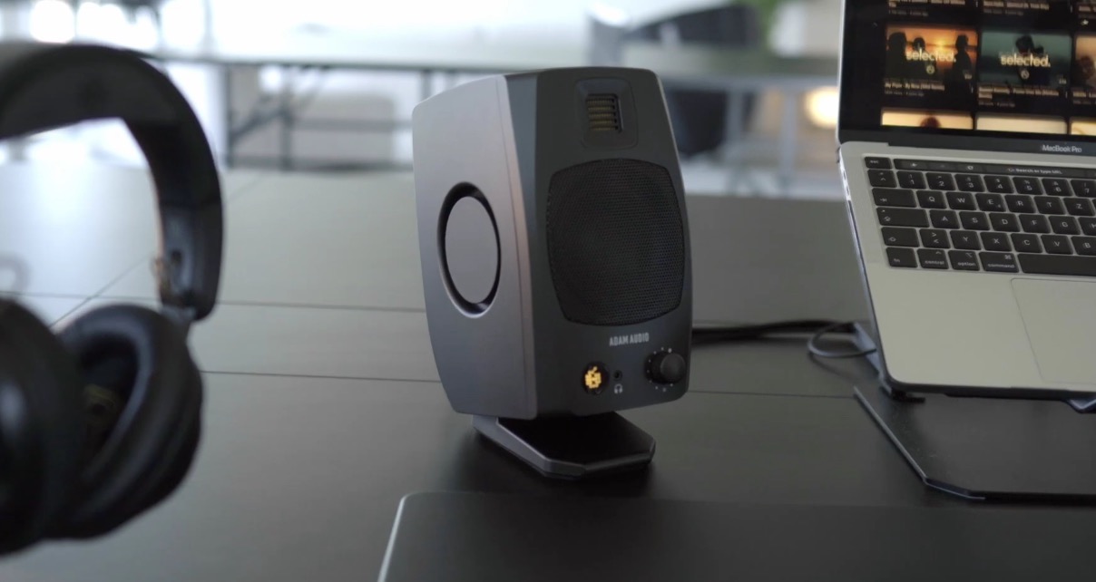 Black headphones, a single Adam Audio D3V speaker with a glowing cyan LED icon, and a silver laptop sit on a black office desk. 