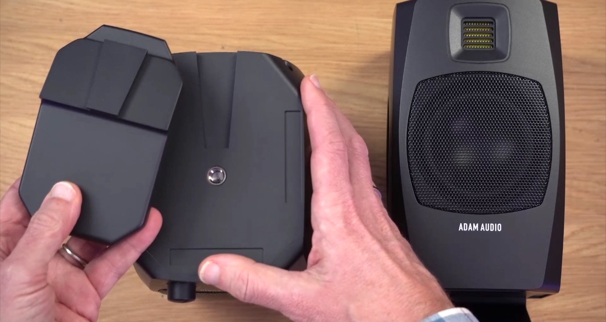 Two black D3V speakers sit on a wooden desk. The one on the left is held by a white man showing both a speaker stand and the thread for mounting. 