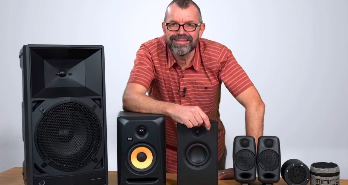 A white, bearded man in his 50s wearing glasses and a red striped polo shirt leans behind 5 different pairs of speakers.