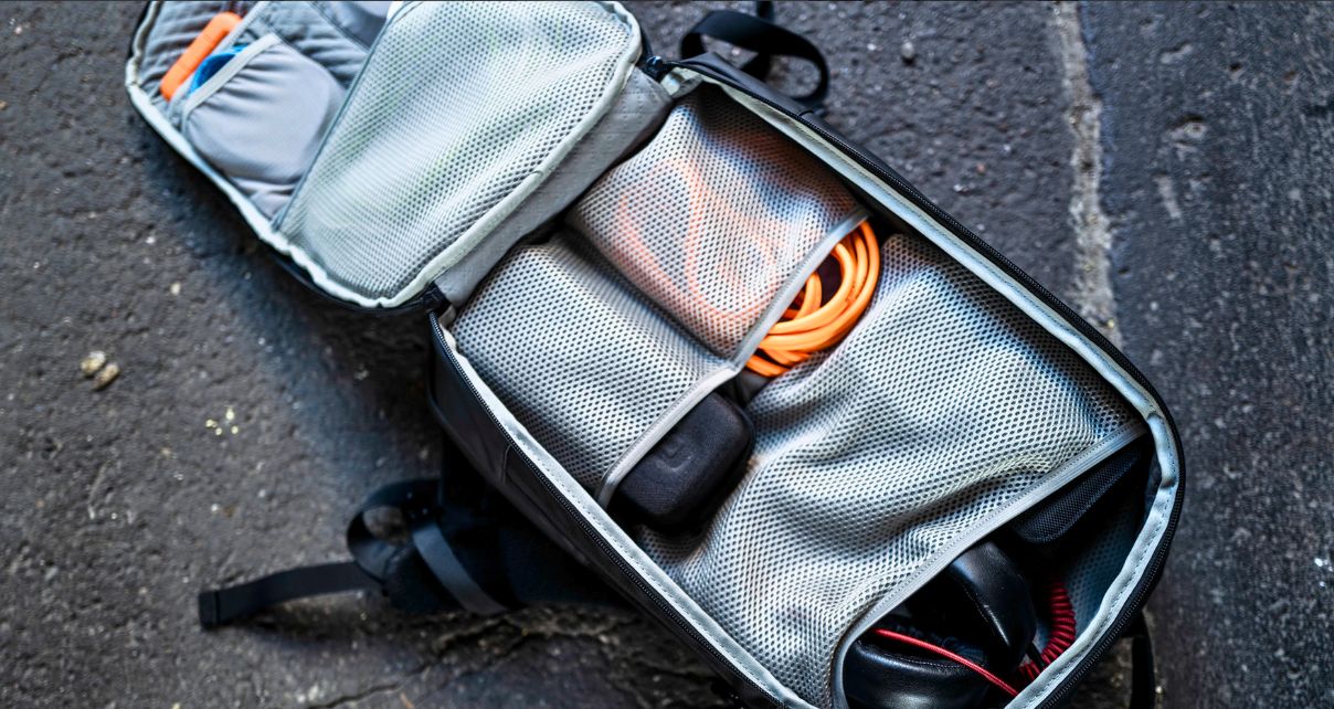 A backpack lays unzipped on asphalt, showing various items inside, including headphones and orange cables.