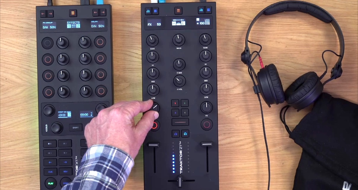 Two black modular Traktor controllers sit next to each other on a wooden desk. A white person with a blue flannel shirt adjusts the controls on one of the units. 
