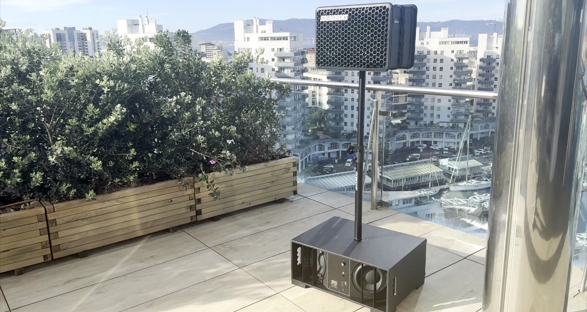 The focal point of this image is a black Soundboks Go portable speaker attached via a black pole to a large Dillinger Labs Death From Below subwoofer on the ground. The setting is a high rise terrace with plants and building in the background. 