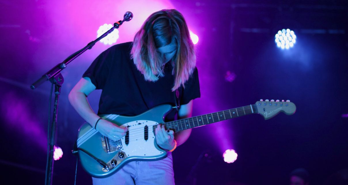 A long-haired guitarist in their 20s plays onstage against a backdrop of purple and pink lighting. 