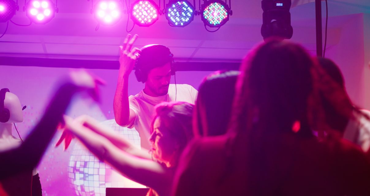 Male DJ wearing a beanie and headphones leads a small party from the stage. In the background are various lights with pink and blue hues, and in the foreground is a handful of people dancing.