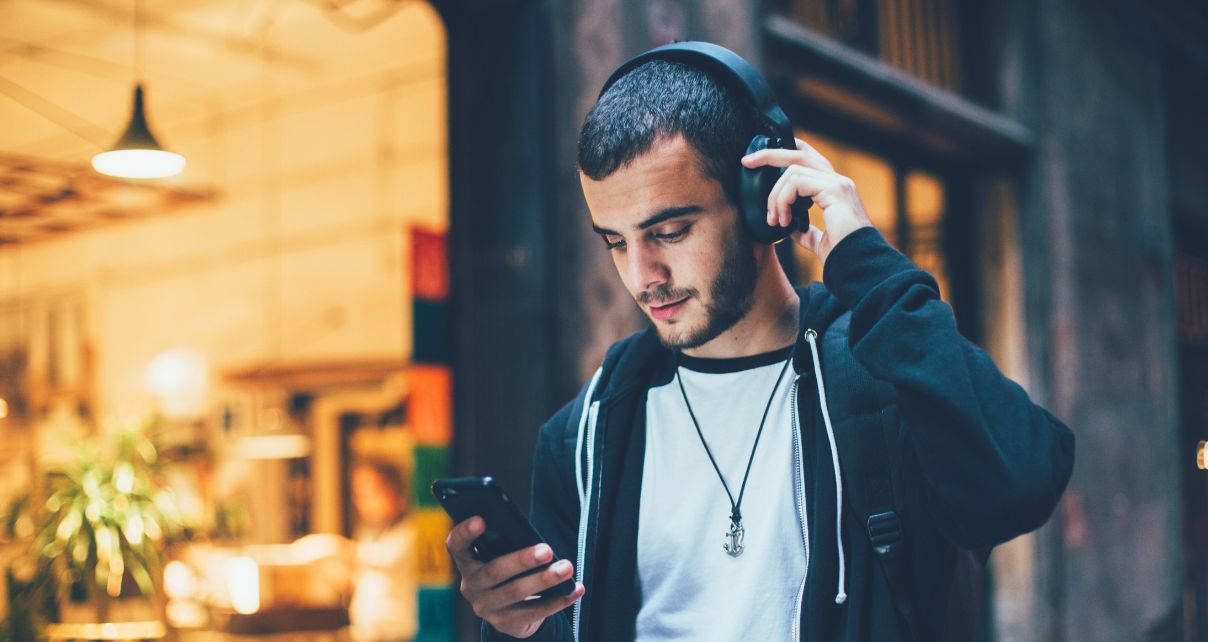 A young male with a dark beard and short hair walks along shops while listening to music and looking at his phone.