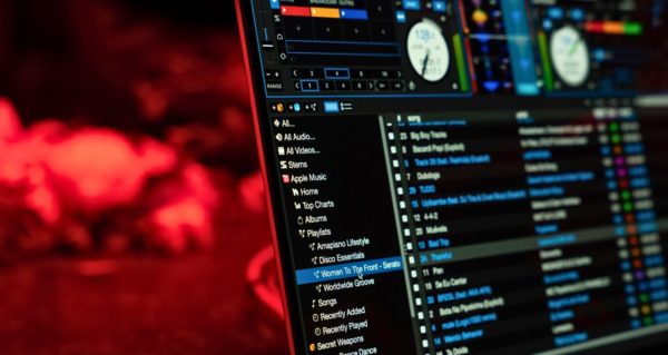 A moody photo with red lighting. In the foreground is a laptop displaying Serato DJ software. Along the left side of the screen is a library tree containing various music playlists. There is one playlist highlighted in blue. The title is "Women To The Front". 