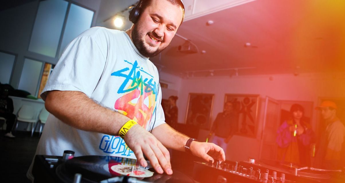 A smiling, bearded white man DJs at an art gallery on turntables. He's wearing black headphones and a white shirt with graffiti writing.