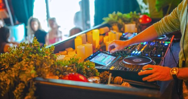 An older white male DJ stands behind a heavily decorated booth. Surrounding the black CDJ set-up in front of him are several plants and candles. The DJ is scrolling through their music library with their right hand, while their left hand rests near the play button. In the background, several people appear to be having lunch.