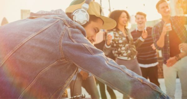 In the foreground, a young male DJ leans over the booth, smiling. He's wearing a hooded jean jacket, tan baseball hat, and silver headphones. In the background several people are dancing, laughing, and drinking. The setting looks like an evening rooftop party.