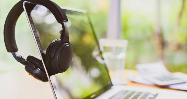 A silver laptop sits on a cozy wooden table. In the daylight, it reflects the surrounding greenery. A pair of black headphones rests over the screen.