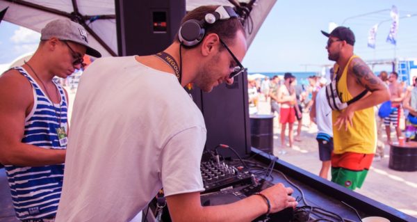 Two DJs stand underneath a tent during a sunny beachside music festival. They both appear to be young white men.
The one on the left is wearing sunglasses, a white and blue striped tank top, a grey baseball hat, and a festival pass lanyard.
The DJ on the right is wearing a plain white shirt, sunglasses, and silver headphones with black flourishes. He's looking down and smiling while selecting the next track on Pioneer DJ CDJs.
In the background is a crowd of people in beachwear enjoying the festival.