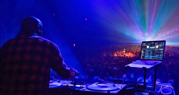 A DJ faces a big crowd in a night club. In front of him are turntables and to the right is a laptop displaying Serato DJ software. 