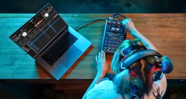 Bird's-eye view of a DJ using the Roland SP-404MKII with a silver laptop running Serato DJ Pro software. Everything is set on a dark wooden table, bathed in turquoise lighting, and the DJ is wearing black V-Moda headphones.