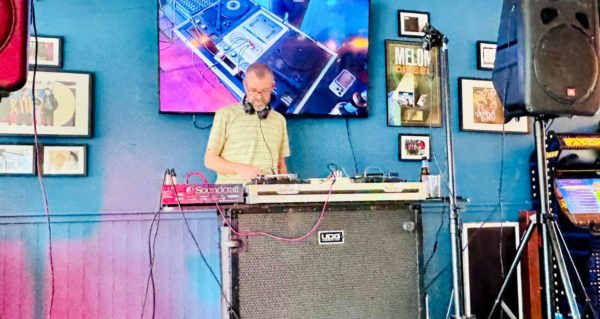 Digital DJ Tips founder Phil Morse stands behind a custom DJ booth. Behind him is a large TV showing the DJ's perspective. There's a large grey and black UDG table holding the gear up, and large speakers mounted on poles on either side of the booth.