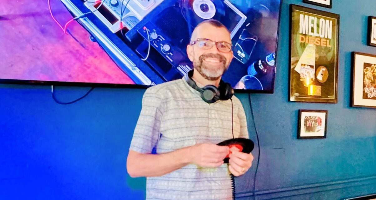 Digital DJ Tips founder Phil Morse smiling in front of a large TV displaying a vinyl DJ set-up. Phil is a white man with glasses and a beard, wearing black headphones around his neck and a light striped t-shirt. He's looking at the camera and holding a seven-inch record with both hands.