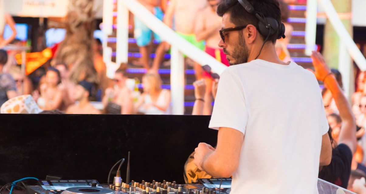 A bearded white male DJ wearing sunglasses and a white T-shirt stands behind a booth. It's a sunny day, and it looks like he's playing to a large crowd at a festival.