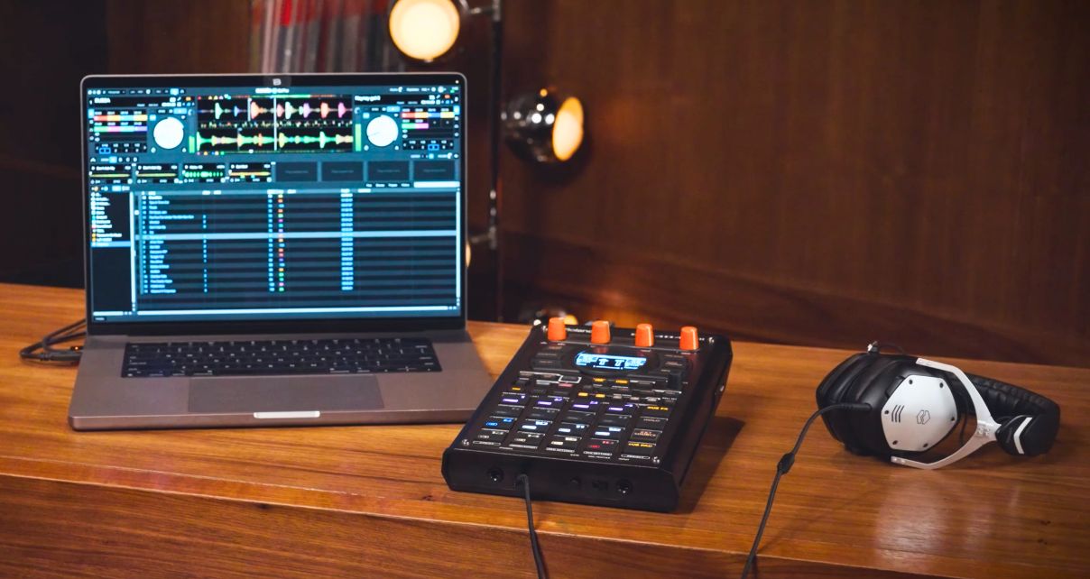 A silver laptop sits on a wooden table next to a Roland SP-404MKII.