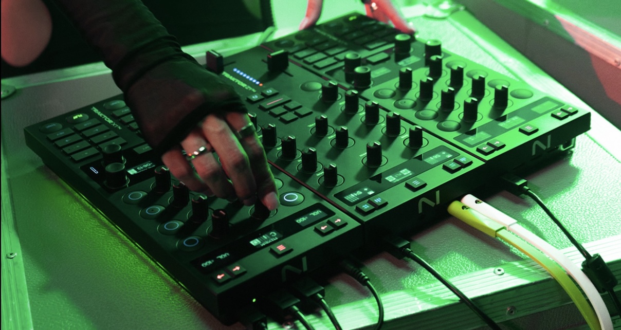 A DJ stands behind three modular Traktor units, tweaking a knob on the mixer. The photo is bathed in green lighting.