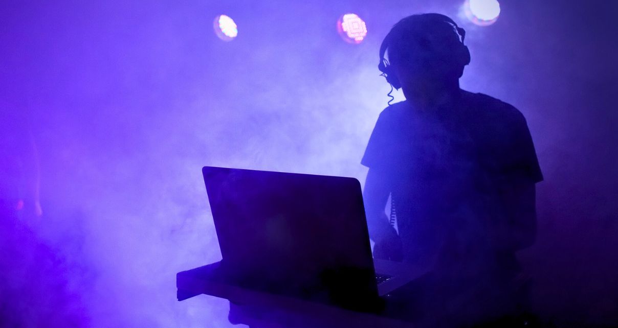 The silhouettze of a DJ stands behind a laptop. Behind the DJ are several stage lights, and the whole scene is bathed in a purple smoke machine haze.
