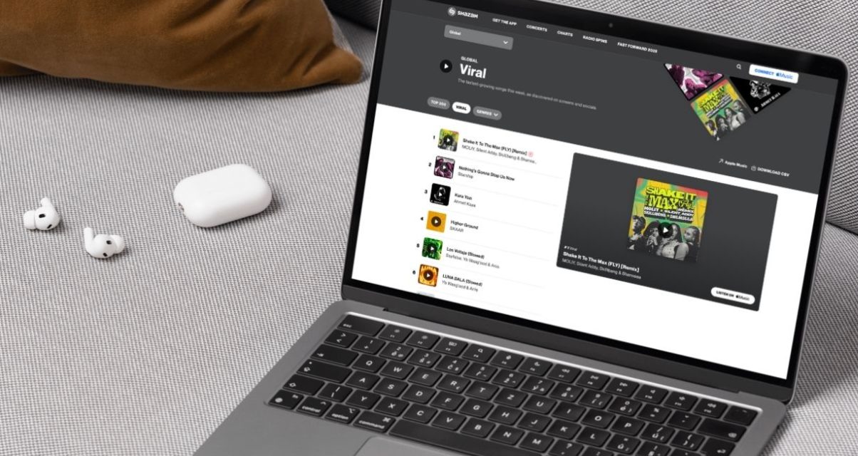 A silver laptop resting on a cream-coloured couch, next to a pair of wireless ear buds. On the laptop screen is Shazam's Viral charts page.