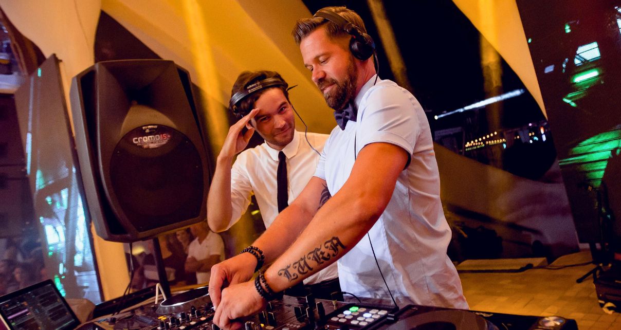Two younger white male DJs stand behind a professional booth. The one on the left is saluting the camera, and the one on the right is tweaking things on the DJ mixer.