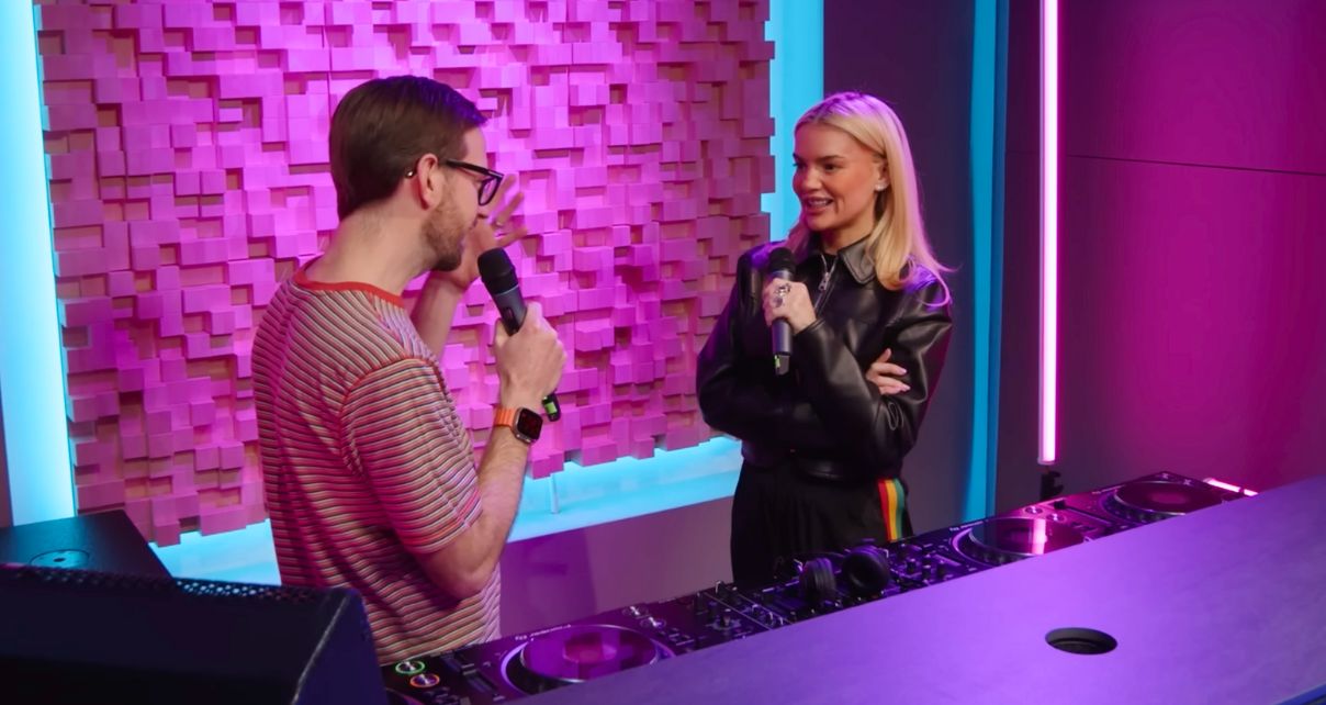 Two people holding microphones stand behind a pro DJ set-up in a bright pink and blue studio. The white man on the left has short brown hair, dark glasses, a short beard, and is wearing a red and white striped t-shirt. On the right is a white woman with blonde hair, wearing a black leather jacket. They are both smiling while talking to each other.