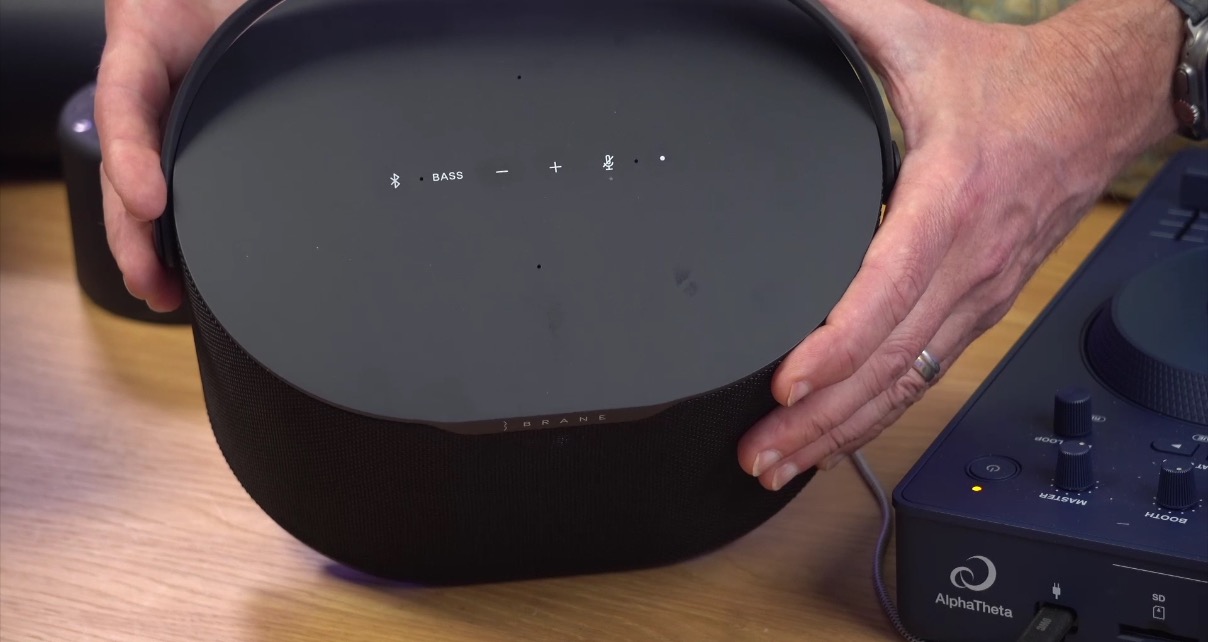 A white person holds a black Brane X speaker with both hands, displaying the controls on the top of the unit