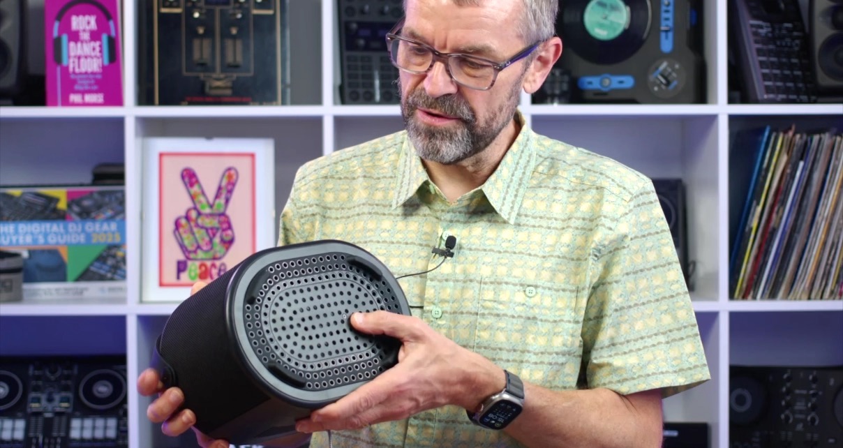 Digital DJ Tips founder Phil Morse holding a black Brane X speaker while standing in front of a studio bookshelf. Phil is white with a greying beard, wearing dark eyeglasses and a light green striped button-up shirt. He's looking down, holding the speaker with both hands, and showing off the bottom subwoofer. 