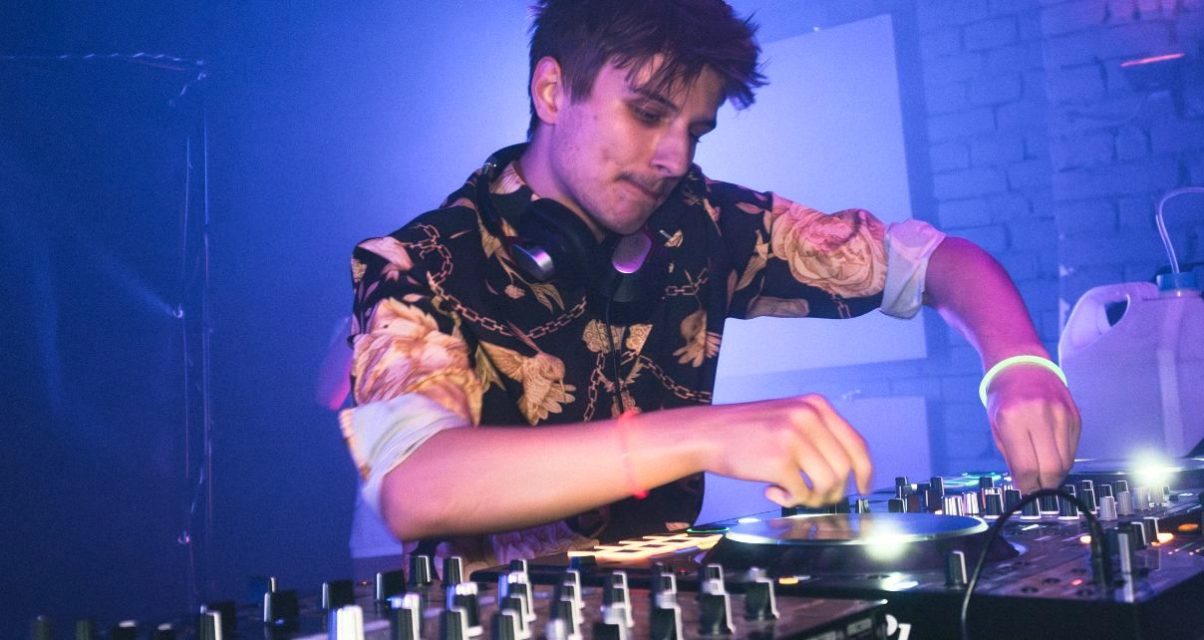 A young male DJ with short brown hair stands behind a professional DJ booth in a nightclub. He's looking down at the gear and tweaking various knobs.