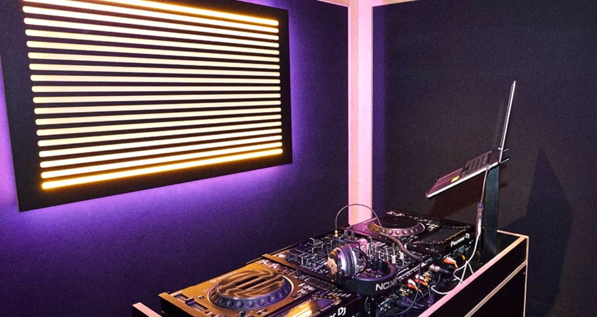 A warmly lit DJ studio, with CDJs, a mixer, headphones, and a laptop.