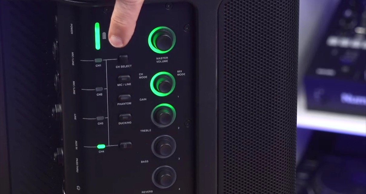 The mixer section on a single black JBL Eon One Compact speaker. The buttons an dknobs have light green LED indicators. At the top you can see a white person points at the full green battery level indicator from offscreen.
