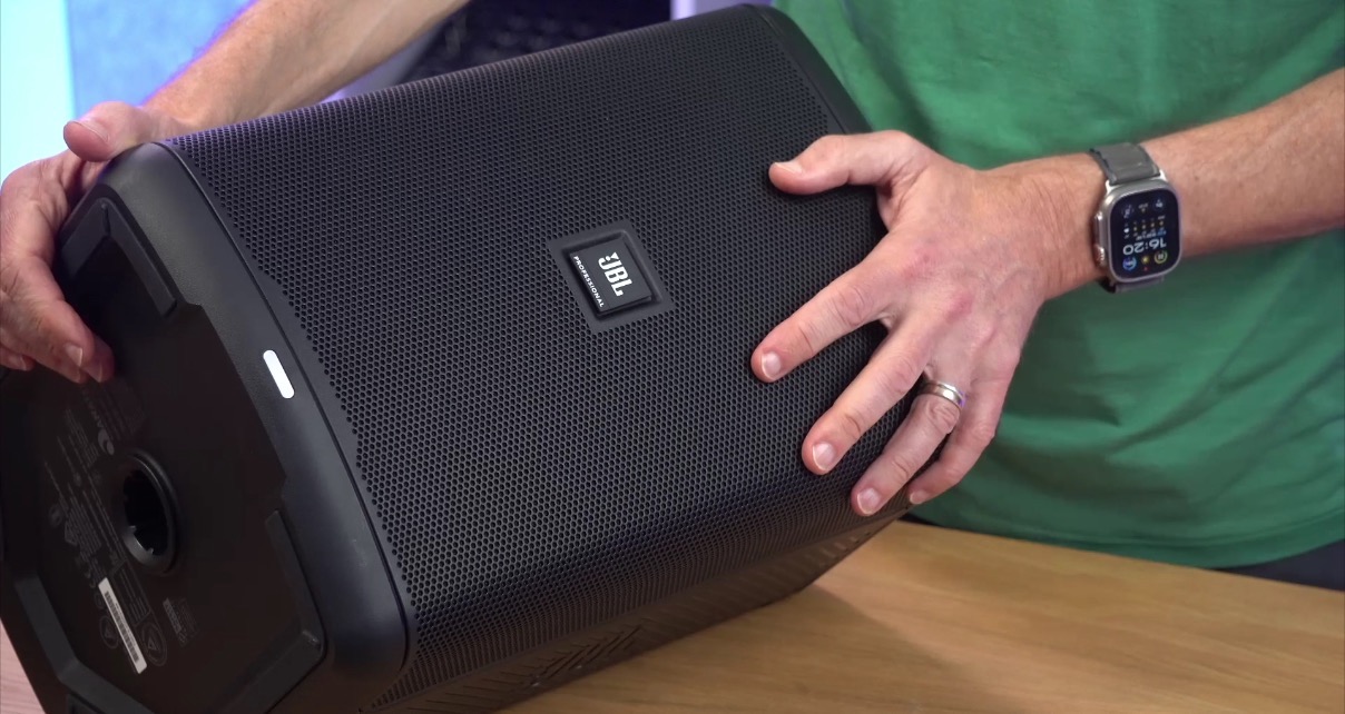 A BLACK JBL Eon One Compact speaker is set on a wooden desk horizontally as a wedge speaker. A white person wearing a watch and light green shirt holds the monitor with both hands.